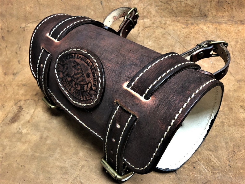 May include: A brown leather arm guard with white stitching and a brass buckle. The arm guard has a circular embossed logo that reads "Owners Special Service" and "Genuine Leather".