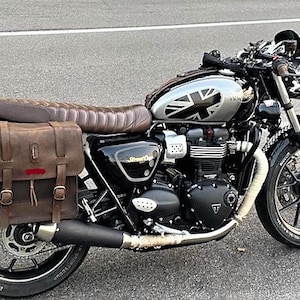 May include: A black motorcycle with a brown leather saddlebag on each side. The motorcycle has a silver gas tank with a Union Jack design. The motorcycle is parked on a gray asphalt surface.