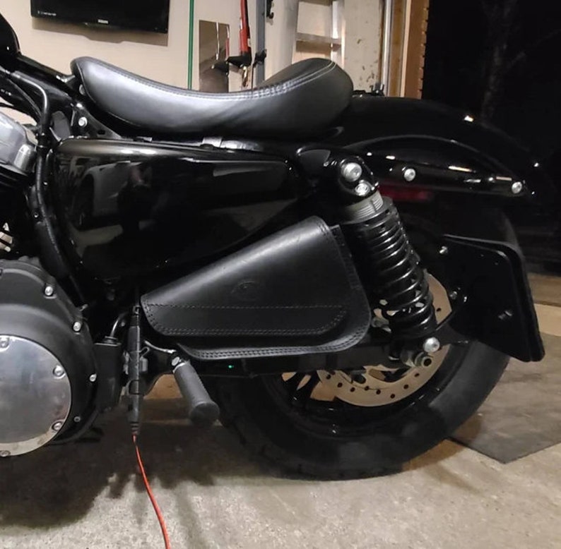 May include: Black motorcycle with a black leather saddlebag attached to the rear fender. The motorcycle has a black seat and a black rear tire.