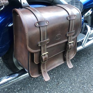 Peut inclure: Une sacoche de moto en cuir marron avec une fermeture à boucle et des détails de couture. Le sac est attaché à une moto bleue.