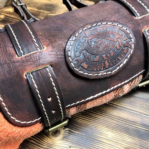 May include: A brown leather motorcycle roll with a white stitched border. The roll has a brown leather strap with a brass buckle. A circular leather patch with white stitching reads "Bikers Special Service PACM Quality Leather Motor Cycles".