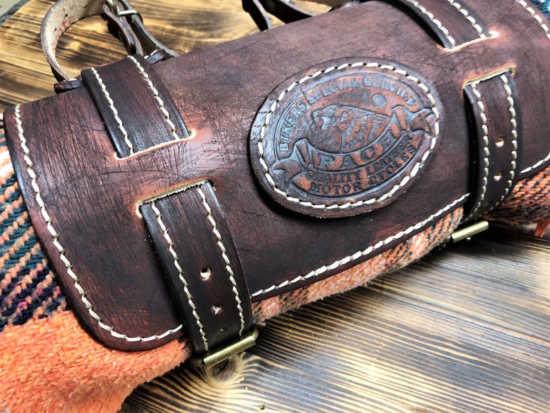 May include: A brown leather motorcycle roll with a white stitched border. The roll has a brown leather strap with a brass buckle. A circular leather patch with white stitching reads "Bikers Special Service PACM Quality Leather Motor Cycles".