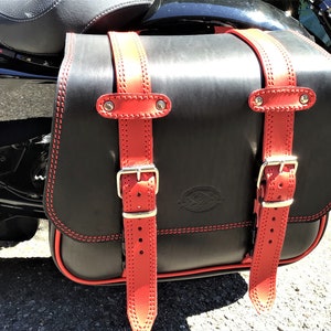 Può includere: Sella da moto in pelle nera con cinturini e fibbie in pelle rossa. La borsa ha un bordo cucito rosso e un piccolo logo sul davanti.