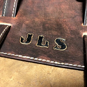 May include: Close-up of a brown leather bag with white stitching. The bag has a personalized inscription that reads "JLS" in black.