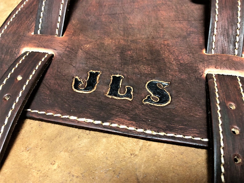 May include: Close-up of a brown leather bag with white stitching. The bag has a personalized inscription that reads "JLS" in black.