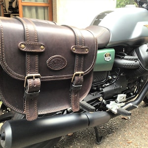 Peut inclure: Une sacoche de selle de moto en cuir marron avec des boucles et des coutures. Le sac est attaché à une moto verte et argentée.