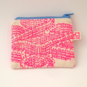 May include: A small, white canvas zippered pouch with a pink city skyline print and a blue zipper. The pouch has a small, white tag with a house icon and the text "BBB" on it.