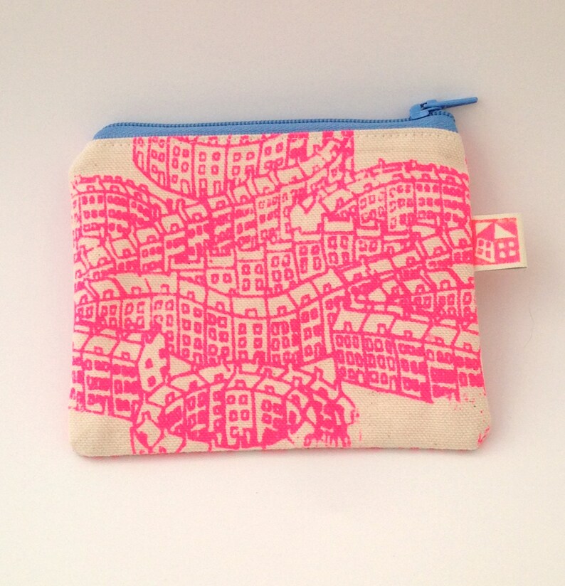 May include: A small, white canvas zippered pouch with a pink city skyline print and a blue zipper. The pouch has a small, white tag with a house icon and the text "BBB" on it.