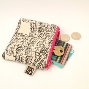 May include: A small, cream-colored zippered pouch with a black and white city map print. The pouch is open and has a pink zipper pull. There are coins and cards inside the pouch.