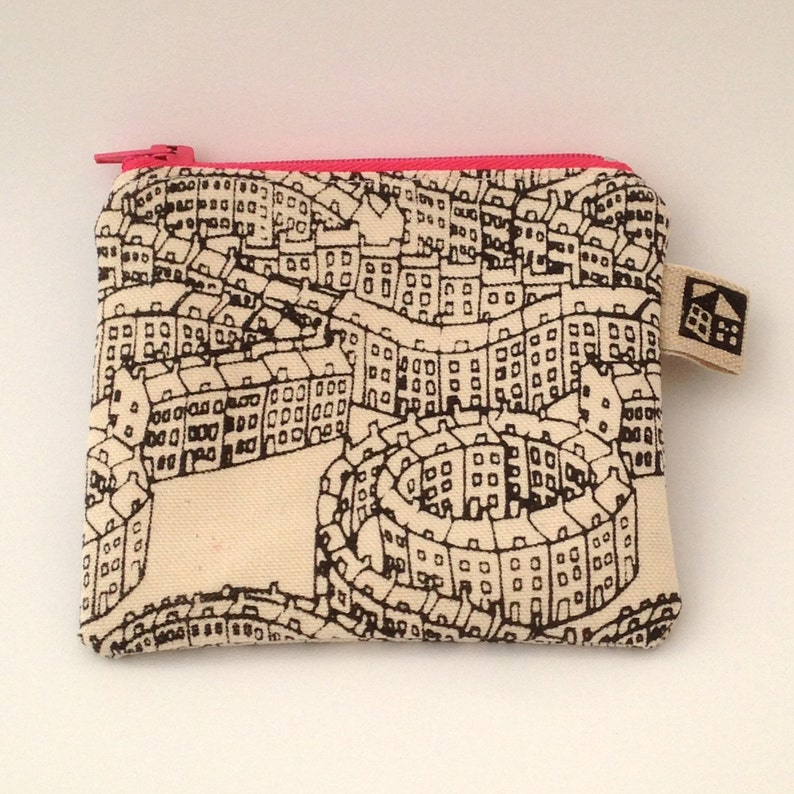 May include: A small zippered pouch with a white canvas background and a black and white print of a city skyline. The zipper is pink.