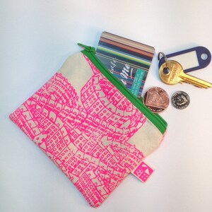 May include: A pink and white fabric pouch with a zipper closure. The pouch is decorated with a pink city map design. The pouch is open and contains a credit card, keys, and coins.