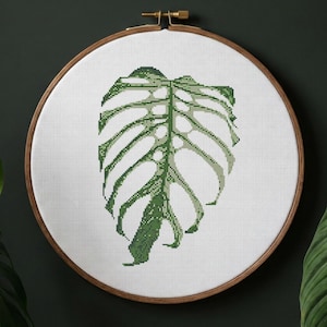 Pode incluir: Um padrão de ponto cruz de uma folha de Monstera, num bastidor de bordado de madeira. A folha é verde com um design detalhado, sobre um fundo branco. O texto "MONSTERA DEVIL MONSTER" e "CROSS STITCH PATTERN" são visíveis.