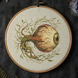 Könnte beinhalten: Kreuzstich-Stickerei eines detaillierten Auges mit Adern und einer braunen Iris, umgeben von grünen Ranken und Blättern. Das Kunstwerk befindet sich in einem hölzernen Stickrahmen, mit dunklem Hintergrund und botanischen Akzenten.