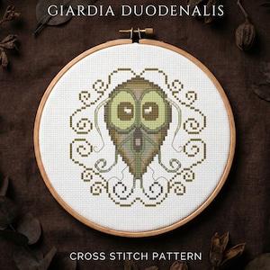 Pode incluir: Um padrão de ponto cruz com um design estilizado de Giardia Duodenalis. O bordado é em tons de verde e castanho, sobre um fundo branco dentro de um bastidor de madeira. O texto "GIARDIA DUODENALIS" e "CROSS STITCH PATTERN" são exibidos.