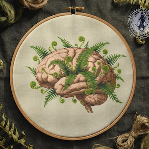 Puede incluir: Bordado de punto de cruz de un cerebro humano con acentos de helechos verdes, en un aro de madera. El cerebro está representado en tonos marrones y beige, con los helechos en verde vibrante. El diseño está sobre una tela color crema.