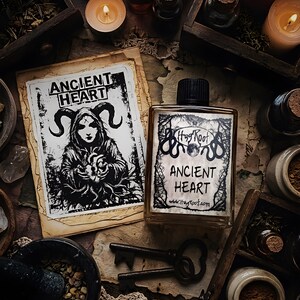 May include: A rectangular glass bottle with a black cap and a label reading ANCIENT HEART and HagRoot.com, accompanied by a paper print featuring a horned figure holding a heart, two metal keys, and a mortar and pestle with herbal contents.