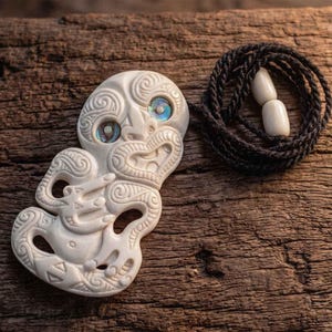 Può includere: Ciondolo color crema scolpito con intricati disegni Maori, con accenti blu iridescenti negli occhi. Una collana con cordino nero con due perline bianche è arrotolata accanto al ciondolo, il tutto su una superficie di legno rustico.