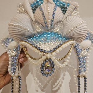 May include: A handcrafted mermaid-themed headpiece featuring seashells, pearls, and blue and clear crystals. The crown has a central teardrop-shaped jewel and dangling pearl strands. The design incorporates white, blue, and gold accents.