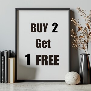 May include: A rectangular black picture frame containing a white print with the bold black text BUY 2 Get 1 FREE. The frame is positioned next to a stack of books, a small speckled stone sphere, and a tall black vase with dried branches.