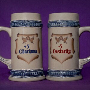 May include: Two beige ceramic beer steins with blue and white trim and handles. One stein reads "+5 Charisma" in blue, and the other reads "-5 Dexterity" in red. Each stein features a crest with two winged creatures.