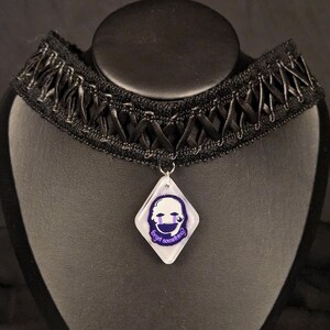 May include: Black choker necklace with a decorative ribbon design. A diamond-shaped pendant hangs from the center, featuring a sad face graphic and the text "forget something?" in purple. The necklace is displayed on a black mannequin.
