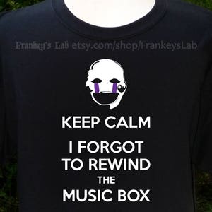 May include: Black t-shirt with a white graphic of a puppet face with purple eyes and the text "Keep Calm I Forgot To Rewind The Music Box".