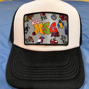 Puede incluir: Gorra de camionero blanca con una malla negra en la parte posterior. La parte delantera de la gorra presenta un parche blanco con un diseño gráfico colorido que incluye el texto "MSG" y varias otras imágenes.