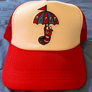 May include: Red and white trucker hat with a red, white and blue worm holding an umbrella with the word "PIPER" on it.