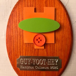 Puede incluir: Placa ovalada de madera teñida de naranja, con un diseño naranja y verde y un botón. Una placa metálica en la parte inferior dice "GUY-YOOT-HEY Hampton Coliseum 2025". La placa está montada sobre una superficie blanca.
