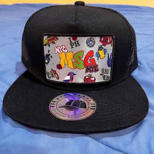 Puede incluir: Gorra de camionero negra y blanca con un parche colorido que presenta el texto "MSG" y varios gráficos, incluyendo un corazón con el texto "I love PH", una dona, una lata de aerosol, un signo de paz y una manzana. La gorra tiene una visera negra y una parte posterior de malla negra. La gorra también tiene una pegatina plateada con el texto "Original Snapbacks" y un diseño de estrella.