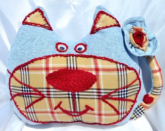 May include: A blue and yellow plaid cat pillow with red embroidered features. The cat has a large, red nose and a stitched smile. The tail is made of blue denim with a red flower.