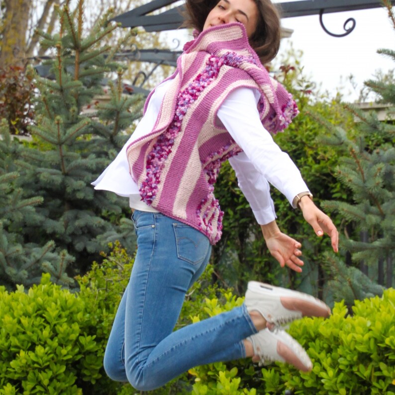 May include: A woman wearing a pink and white striped crocheted vest with a hood. The vest has a textured, fluffy look. She is wearing blue jeans and white sneakers.