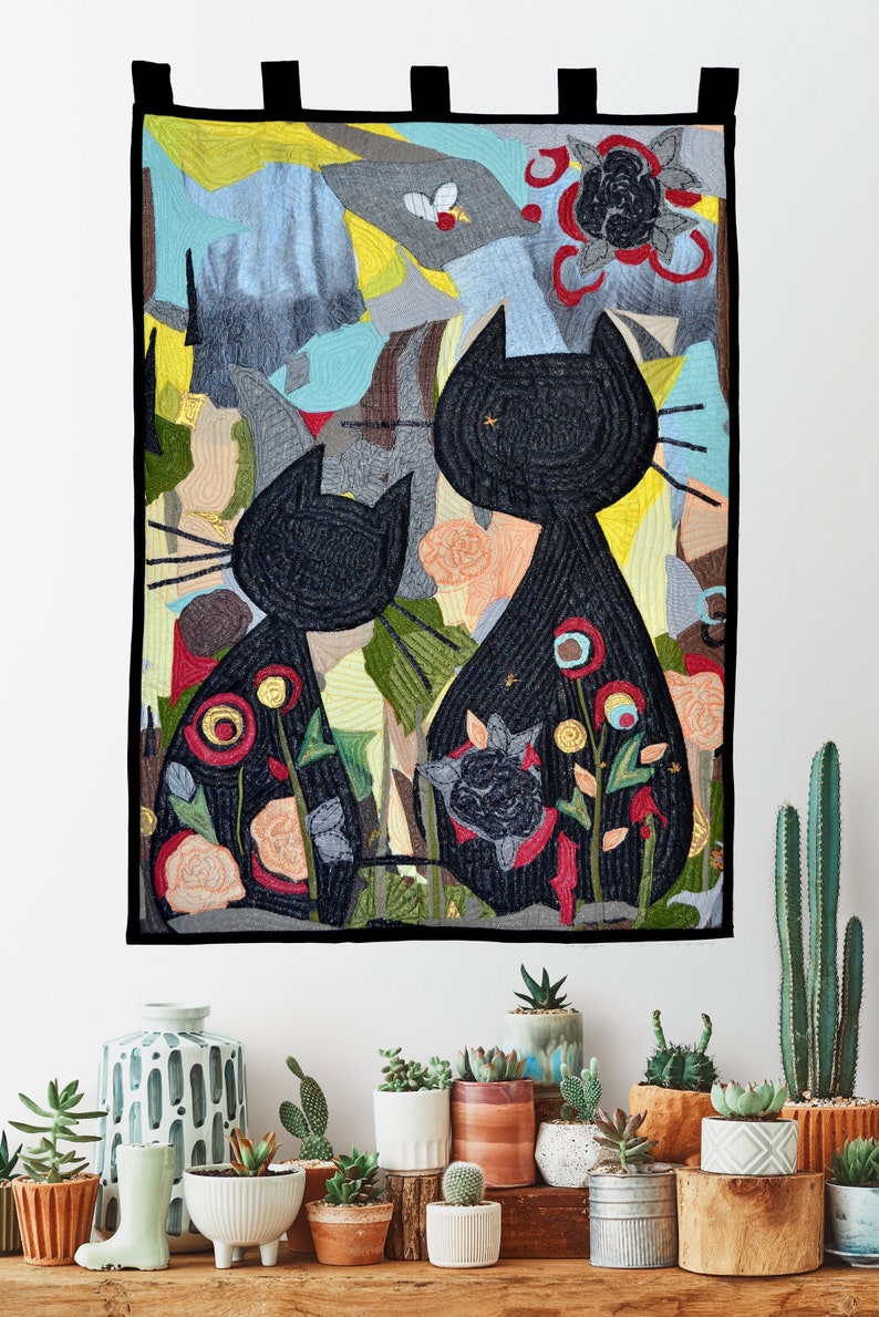Pode incluir: Uma tape&ccedil;aria de parede preta com duas silhuetas estilizadas de gatos pretos com padr&otilde;es florais. O fundo &eacute; um patchwork colorido de azuis, verdes, amarelos e cinzas. A obra de arte &eacute; emoldurada com uma borda preta.