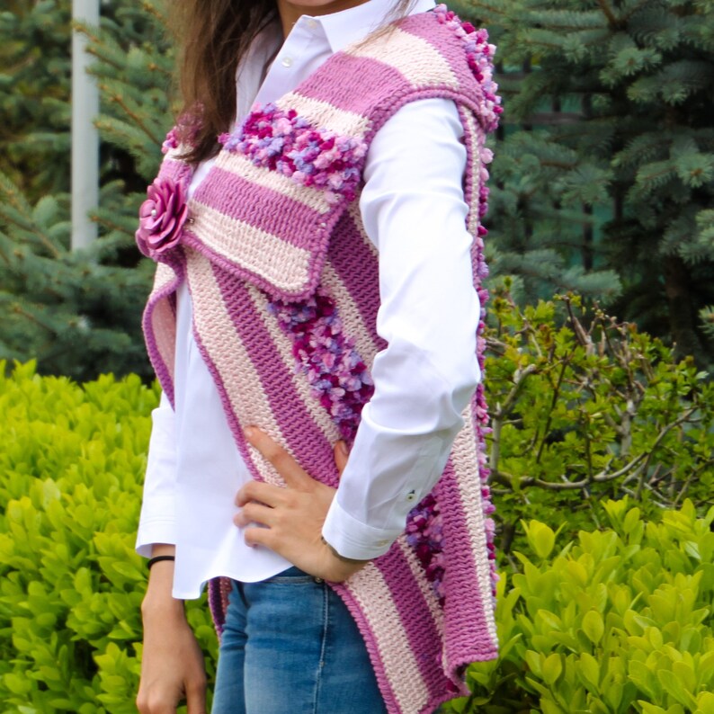 May include: A pink and white striped crochet vest with a flower embellishment. The vest has a long, flowing design and is made with a soft, textured yarn.