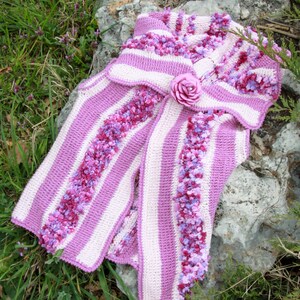 May include: A pink and white striped crocheted vest with a ruffled collar and a pink flower brooch.