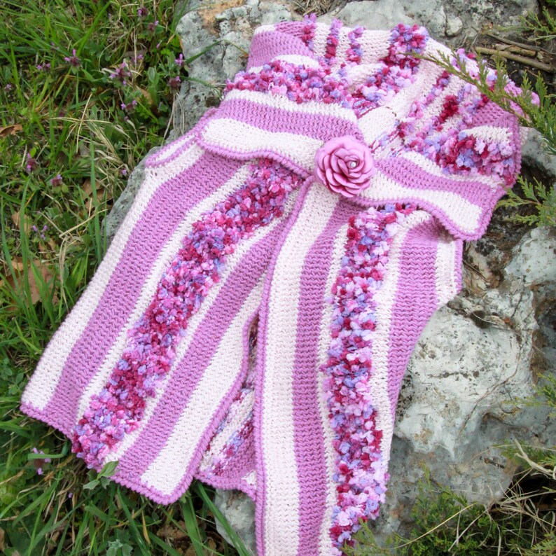 May include: A pink and white striped crocheted vest with a ruffled collar and a pink flower brooch.
