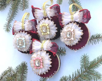 May include: A group of burgundy velvet Christmas baubles. Each bauble has a white tulle flower with a central jewel, a white and gold bow, and a gold hanging loop. The baubles are decorated with small pearls.