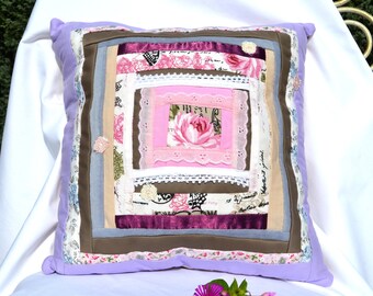 May include: A square pillow with a patchwork design featuring various shades of brown, pink, and white fabric. The pillow has a lavender colored border and is decorated with floral patterns and lace.