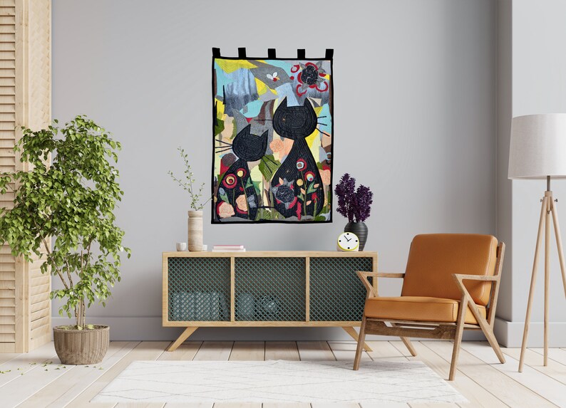 Pode incluir: Uma tape&ccedil;aria decorativa de parede com duas silhuetas de gatos pretos com padr&otilde;es florais e abstratos. A obra de arte est&aacute; sobre um fundo colorido com uma borda preta. &Eacute; exibida acima de um arm&aacute;rio de madeira e uma cadeira de couro.