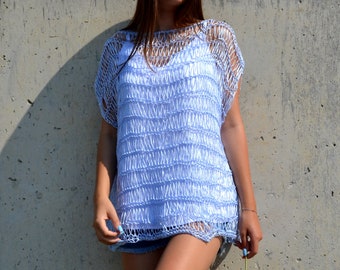 May include: A light blue crochet top with a textured design. The top has short sleeves and a loose fit. The model is wearing the top with denim shorts. The background is a gray concrete wall.