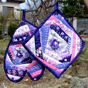 Op de afbeelding: Twee paarse en roze gequilte ovenwanten met bloemenborduurwerk en satijnen lint accenten.