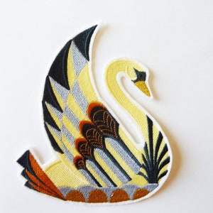Peut inclure: Un cygne jaune avec des ailes noires et grises et des accents marrons. Le cygne est brodé sur un tissu blanc.