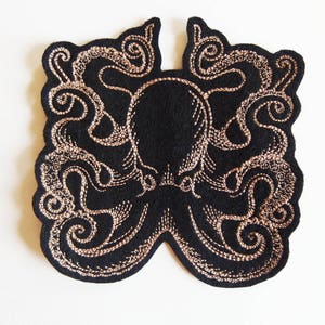 Peut inclure: Un patch brodé noir représentant un poulpe avec du fil doré. Le poulpe a huit tentacules avec des détails complexes.