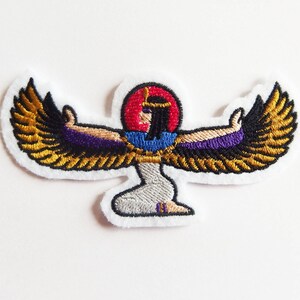 Op de afbeelding: Geborduurde patch met een Egyptische godheid met uitgespreide vleugels. De patch heeft een witte rand, met de godheid afgebeeld in goud, paars, zwart, rood en blauw draad. Het ontwerp doet denken aan oude Egyptische kunst.