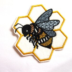 May include: Embroidered patch depicting a bee with black, yellow, and blue detailing, set on a white honeycomb shape with a gold border. Ideal for customising clothing or accessories.