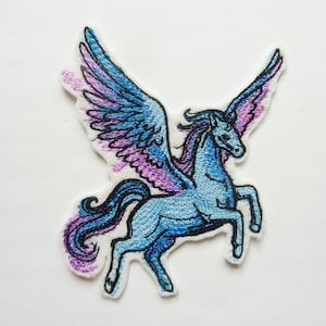 Könnte beinhalten: Gestickter Aufnäher mit einem blauen Pegasus mit lila und blauen Flügeln. Das Fabelwesen hat eine fließende Mähne und einen Schweif und steht auf seinen Hinterbeinen. Der Aufnäher befindet sich auf weißem Hintergrund.