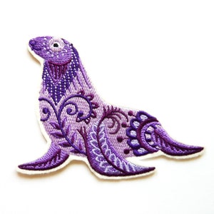 Könnte beinhalten: Gestickter Aufnäher mit einem lila Seehund-Design mit komplizierten Blumen- und Wirbelmustern. Der Seehund ist im Profil dargestellt, mit einem weißen Rand. Der Aufnäher befindet sich auf weißem Hintergrund.