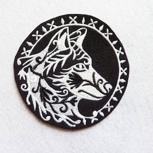 May include: Black and white embroidered patch featuring a wolf's head with intricate floral designs. The wolf's head is surrounded by a circular border with a repeating pattern.