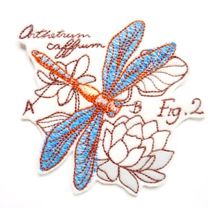 Peut inclure: Un écusson brodé représentant une libellule aux ailes bleu vif et au corps orange, entourée d'illustrations florales aux contours marron. Le texte indique Orthetrum caffrum, A, B et Fig. 2 en fil marron sur un fond en tissu blanc.