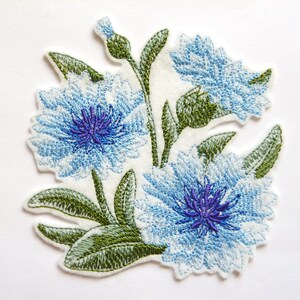 Pode incluir: Um emblema bordado apresentando duas grandes flores de centáurea azul-claras com centros roxo-escuros, acompanhadas por folhas e caules verdes sobre um fundo de tecido branco. O design inclui costuras detalhadas que criam textura.